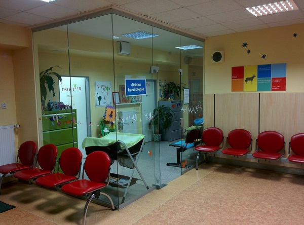 Čekárna s červenými židlemi a prosklenou stěnou ordinace s cedulí „dětská kardiologie“